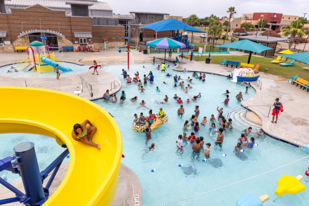 Outdoor Leisure pool - young girl going down a big yellow slide, a large group of people below playing in a large pool with ice cream themed and shaped floats
