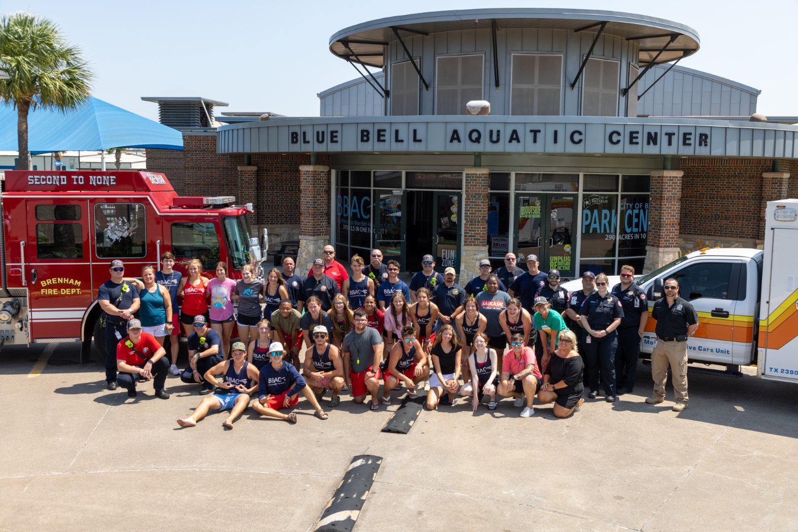 group picture of firefighters, lifeguards, and ems staff in front of a fire truck and ambulance in front of the aquatic center building